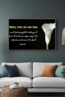 Framed wall art featuring Isaiah 43:19 with the text “Behold, I will do a new thing… I will even make a way in the wilderness, and rivers in the desert” displayed in white and yellow on a black background, alongside a single elegant white calla lily. The artwork is mounted above a modern white sofa with throw pillows.