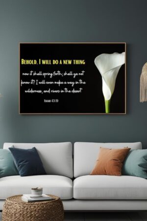 Framed wall art featuring Isaiah 43:19 with the text “Behold, I will do a new thing… I will even make a way in the wilderness, and rivers in the desert” displayed in white and yellow on a black background, alongside a single elegant white calla lily. The artwork is mounted above a modern white sofa with throw pillows.
